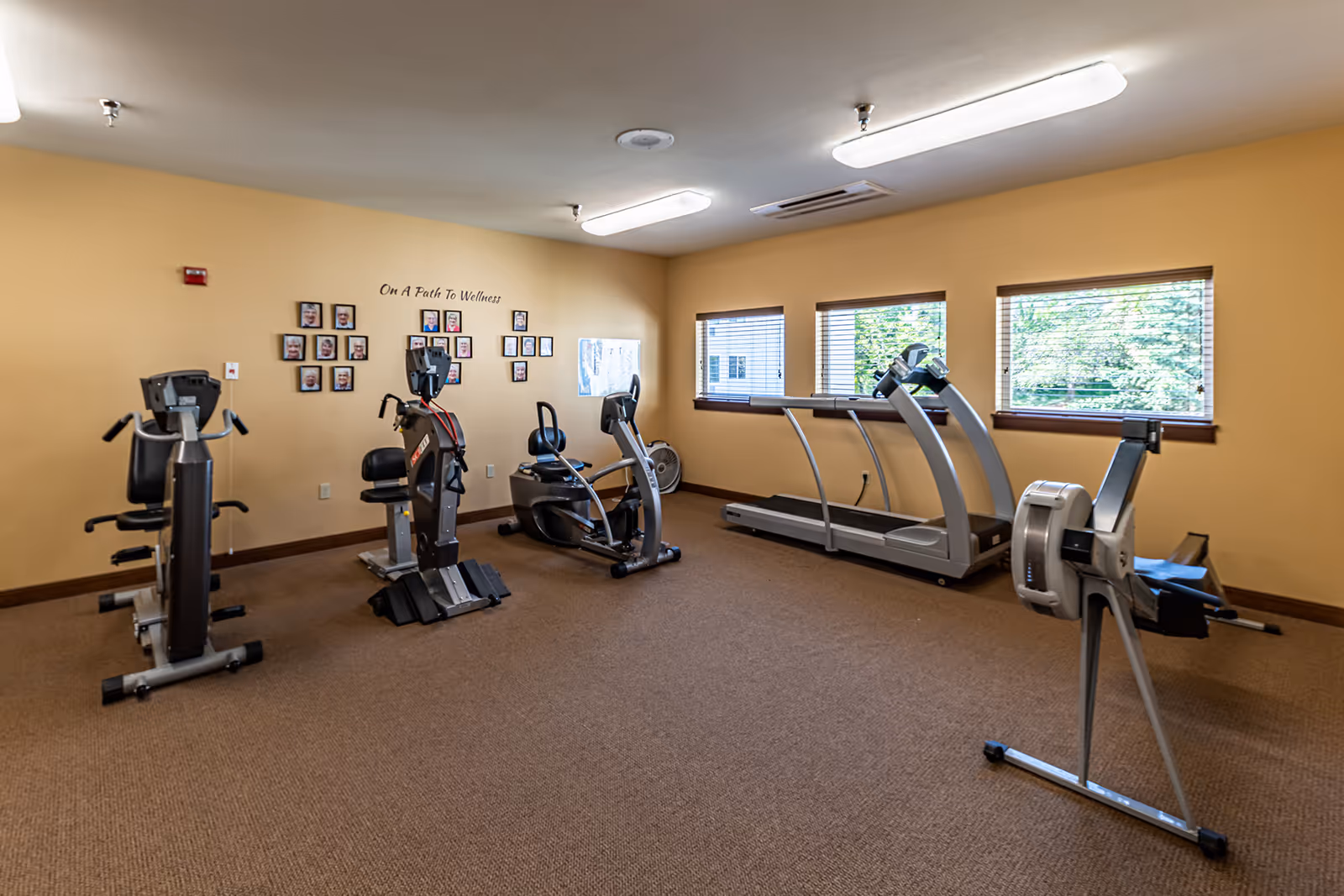 A fitness room with exercise equipment including two recumbent bikes, a treadmill, and a rowing machine. The room has beige walls and carpeted floor, with three windows letting in natural light. On one wall, there are framed photos and the phrase 'On A Path To Wellness' displayed.