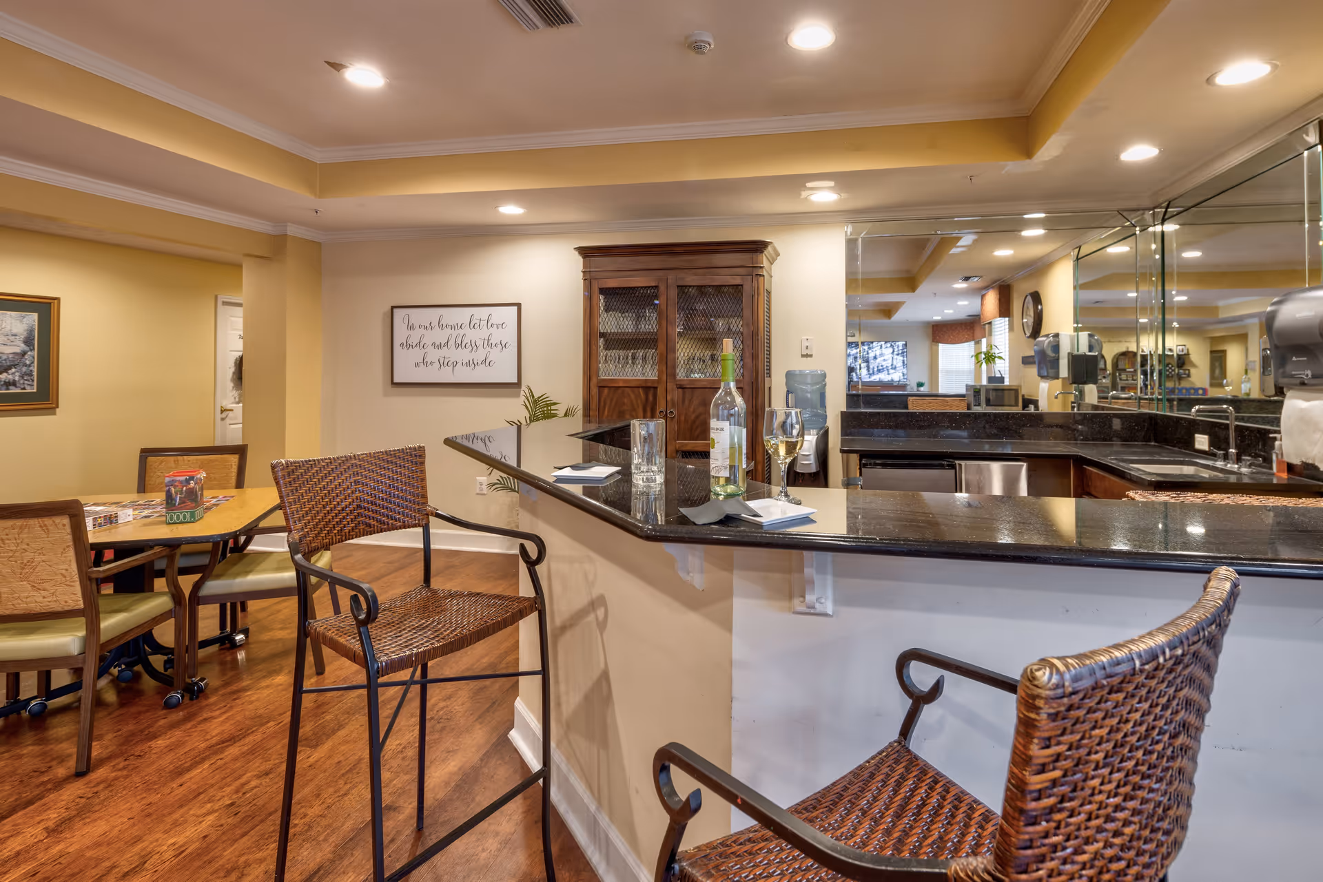 Interior view of a common area in a senior living facility featuring a bar counter with two wicker bar stools, a bottle of white wine, a wine glass, and a water glass on the counter. Behind the counter is a sink, a microwave, and a water dispenser. To the left, there is a table with chairs and a puzzle on it. The walls are painted beige with framed artwork and a decorative sign that reads 'In our home let love abide and bless those who stop inside.' The floor is wooden, and the ceiling has recessed lighting.