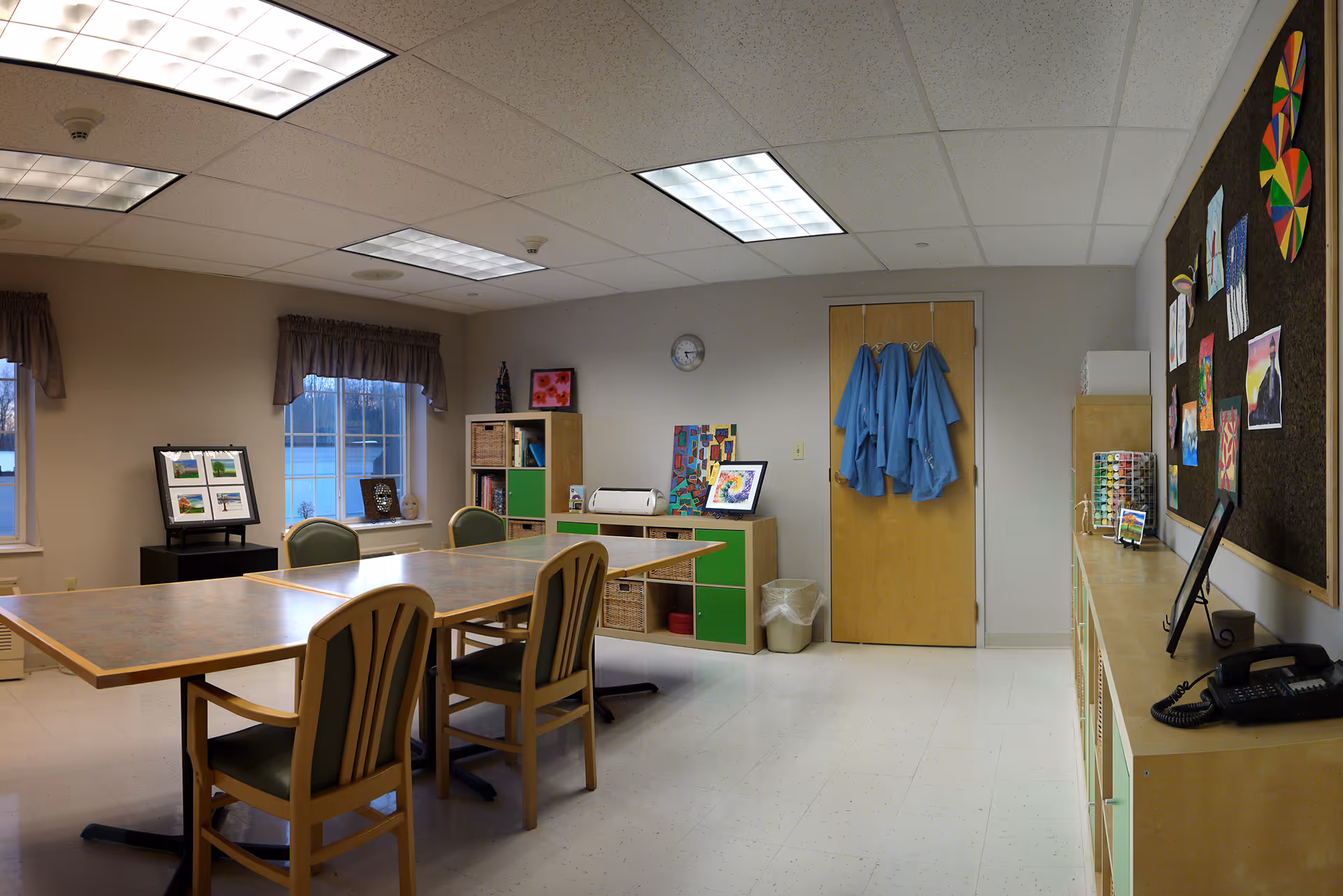 A well-lit activity room with a large table surrounded by chairs. The room has windows with brown curtains, a bulletin board displaying colorful artwork, shelves with storage baskets, and blue aprons hanging on a door. The floor is tiled and the ceiling has fluorescent lights.