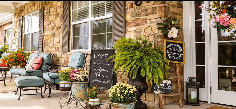 A cozy outdoor patio area with cushioned chairs and ottomans in a soft blue color, surrounded by various potted plants and flowers. There are decorative signs with welcoming messages such as 'HOME is where your story begins. Welcome Home' and 'Welcome Home' displayed near the entrance of a stone building with large windows and a glass door.