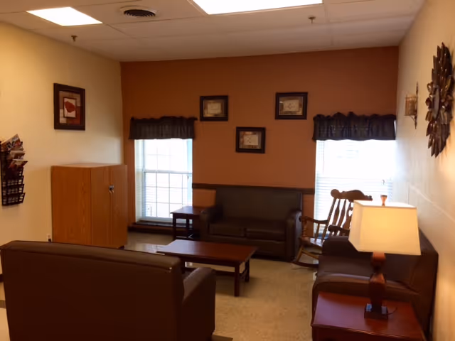 A cozy living room with two brown sofas, a wooden rocking chair, a coffee table, two side tables, a lamp, and framed pictures on the walls. Two windows with dark valances allow natural light into the room.