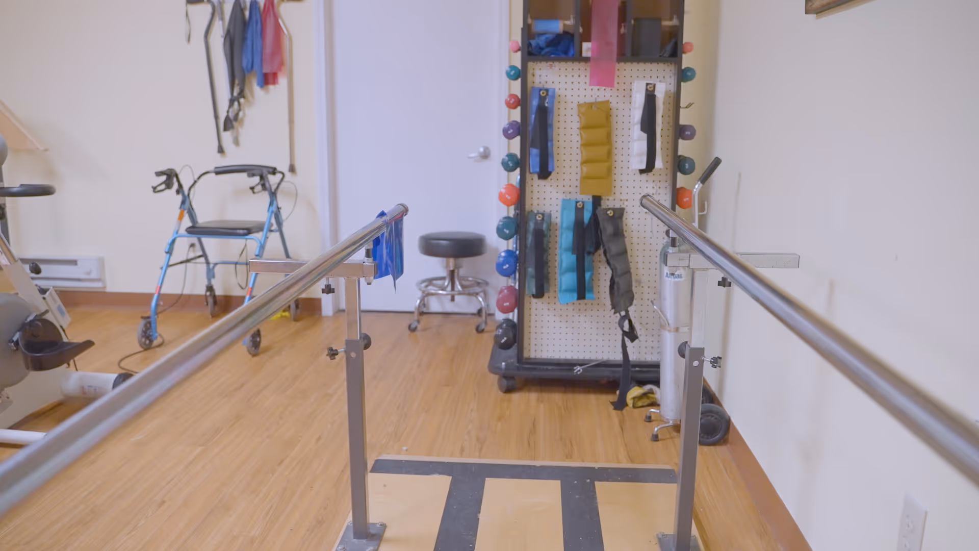 Physical therapy room with parallel bars for walking exercises, a walker, exercise bike, resistance bands, dumbbells, and other rehabilitation equipment on a wooden floor.