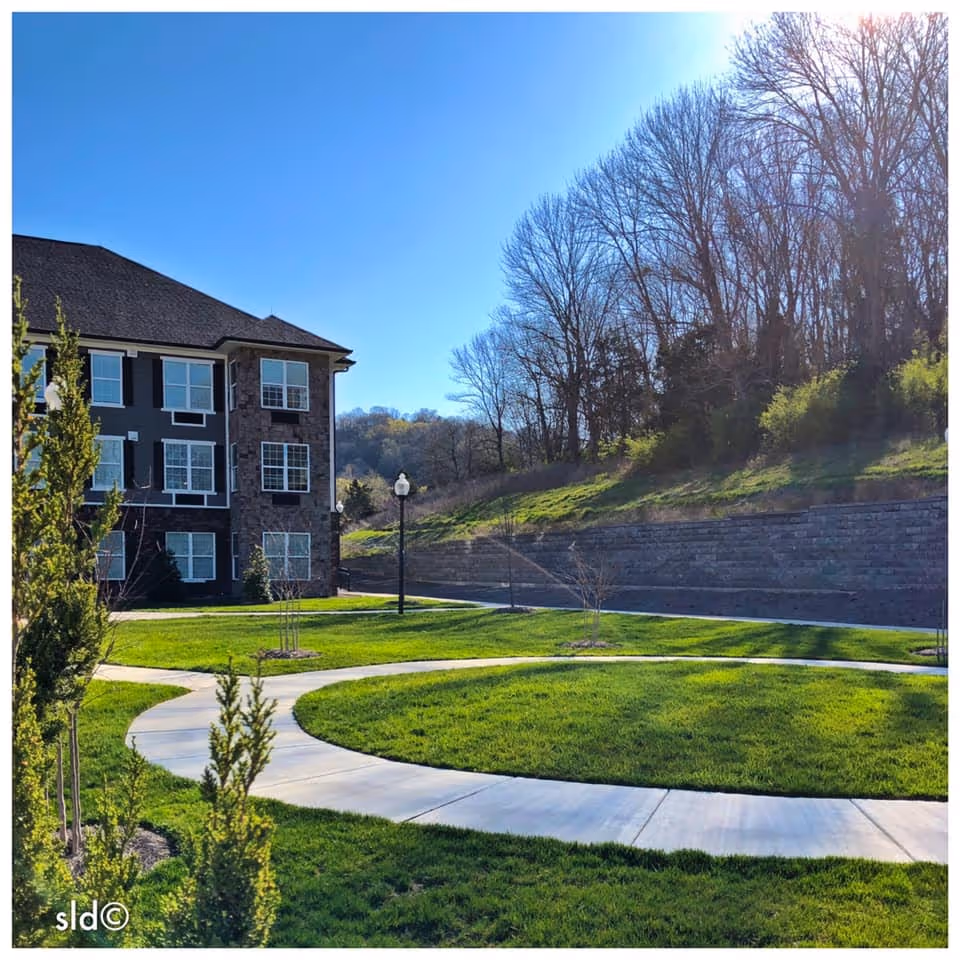 A sunny outdoor scene at Canterfield Senior Living showing a curved concrete walkway surrounded by well-maintained green grass and small trees. To the left is a multi-story building with large windows and a stone facade. In the background, there are leafless trees and a retaining wall under a clear blue sky.