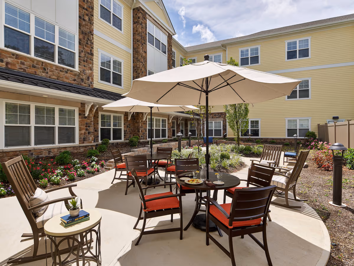 Outdoor patio area at Tappan assisted living nursing home with round tables, chairs with red cushions, large beige umbrellas, and a small side table with books and a plant. The patio is surrounded by a garden with flowers and shrubs, and the building exterior features yellow siding and stone accents.