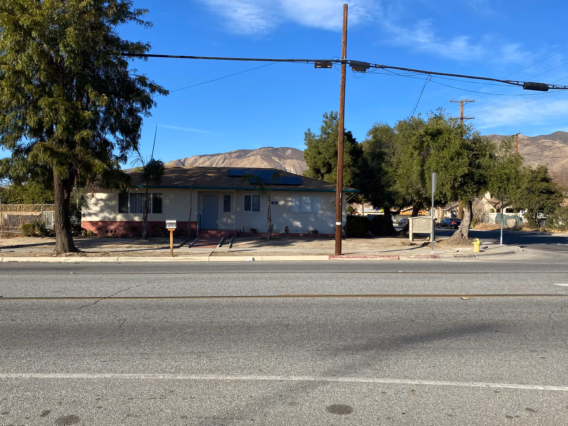 Single-story building with a light-colored exterior and a dark roof, situated behind a sidewalk and a street with visible power lines overhead. There are several trees around the building and mountains in the background under a clear blue sky.