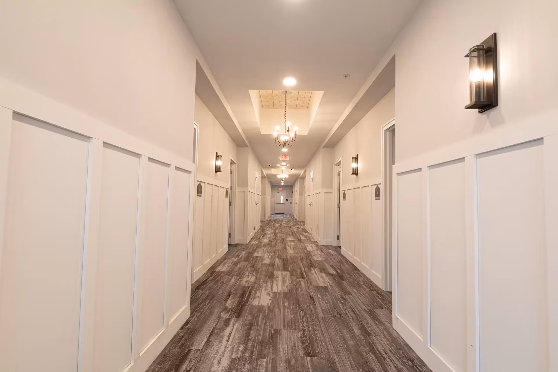 A long, well-lit hallway in a senior living facility with white paneled walls, multiple doors on both sides, modern wall sconces, and chandeliers hanging from the ceiling. The floor has a wood-like pattern.