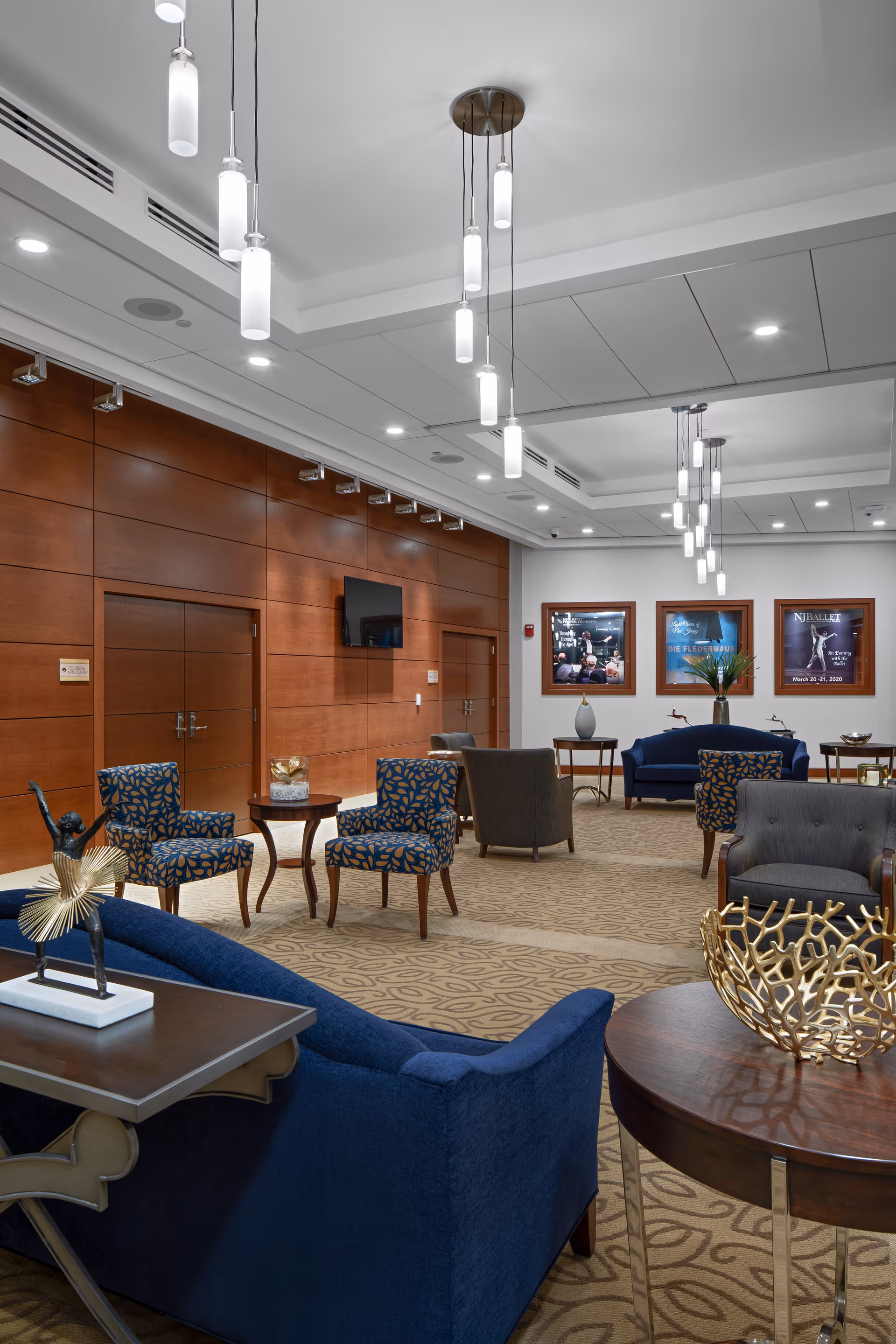 A spacious and well-lit lounge area with blue and patterned armchairs, a blue sofa, wooden tables, and modern pendant lights hanging from the ceiling. The walls are paneled with wood, and there are framed posters on the far wall. The carpet has a subtle leaf pattern.