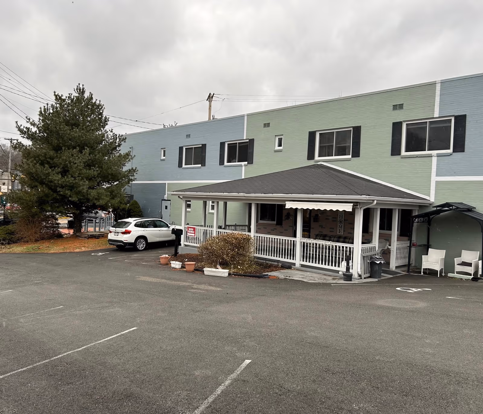 Exterior view of a two-story building with a covered porch area, several windows with black shutters, a white car parked in the lot, and some outdoor seating. The building has a green and blue painted facade with white trim. The sky is cloudy.
