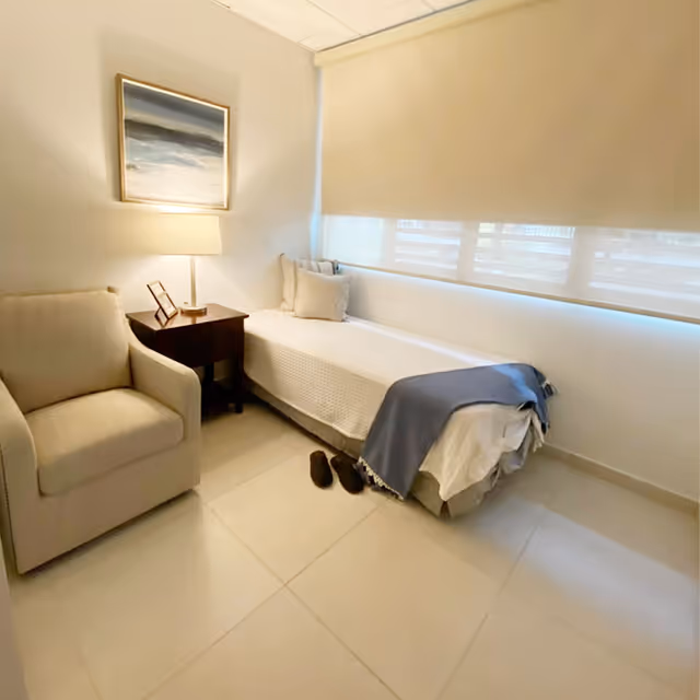 Small bedroom with a single bed by a window, an armchair, a nightstand with a lamp, and slippers on the floor.