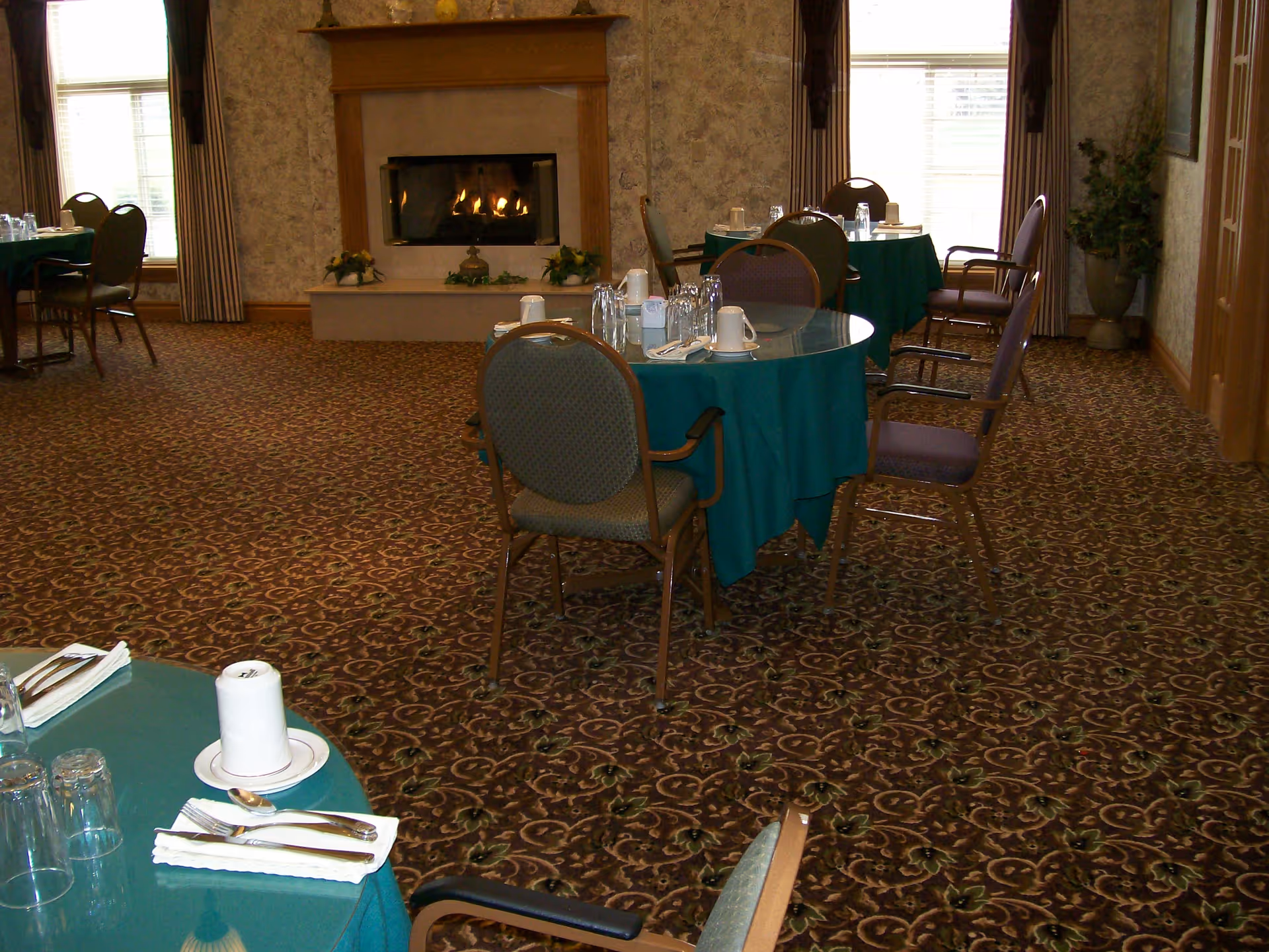 Dining room with round tables covered in green tablecloths, set with cups, glasses, and silverware. Several chairs are arranged around the tables. A lit fireplace with a wooden mantel is visible against the far wall, flanked by windows with curtains. The room has patterned carpet and wallpaper, and a potted plant is in the corner.