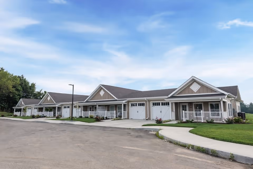 Exterior view of a single-story residential building with multiple units, each featuring a garage and a small porch with white railings. The building is surrounded by a paved driveway and well-maintained green lawns under a partly cloudy blue sky.
