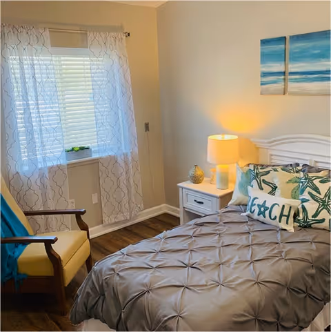 A cozy bedroom with a neatly made bed featuring a gray comforter and decorative pillows, including one with the word 'BEACH'. Next to the bed is a white nightstand with a lit lamp and decorative items. A window with sheer patterned curtains lets in natural light. A wooden armchair with a yellow cushion and a blue throw is positioned near the window. The walls are light-colored with two blue ocean-themed paintings above the bed.