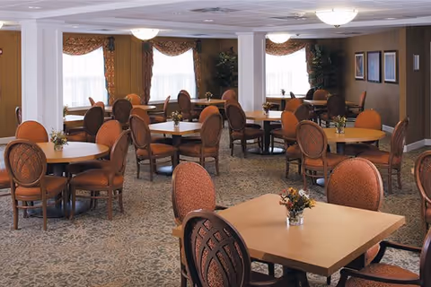 A spacious dining room with multiple round and square tables surrounded by cushioned wooden chairs. Each table has a small floral centerpiece. The room has large windows with decorative curtains, carpeted floors, and warm lighting from ceiling fixtures. There are framed pictures on the walls and some potted plants in the corners.