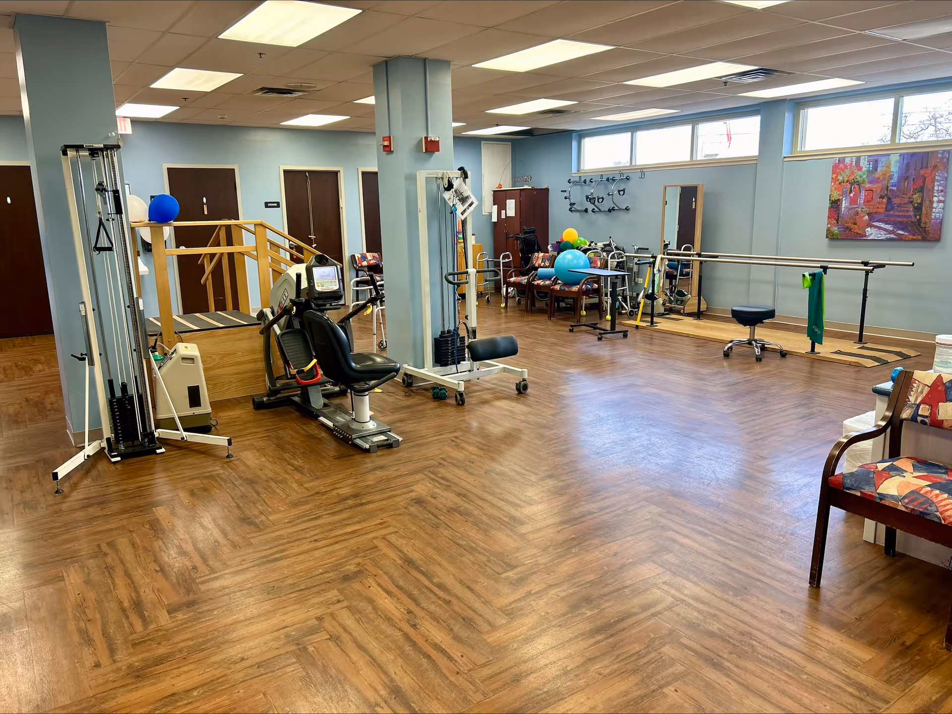 A spacious rehabilitation or physical therapy room with exercise equipment, parallel bars for walking practice, chairs, therapy balls, and a wooden floor. The room has light blue walls, several doors, large windows near the ceiling, and a colorful painting on the wall.