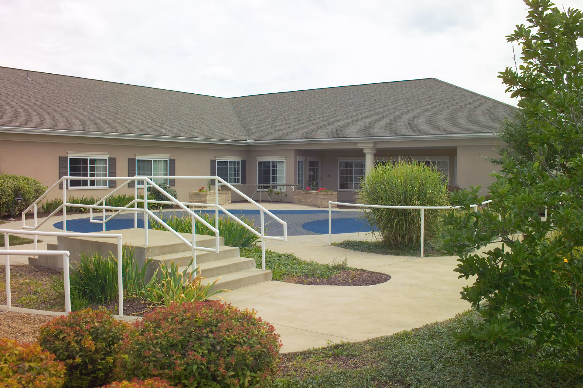 Exterior view of a single-story building with beige walls and a gray roof, featuring multiple windows and a covered entrance. There is a paved walkway with handrails, steps, and landscaped greenery including bushes and plants in the foreground.