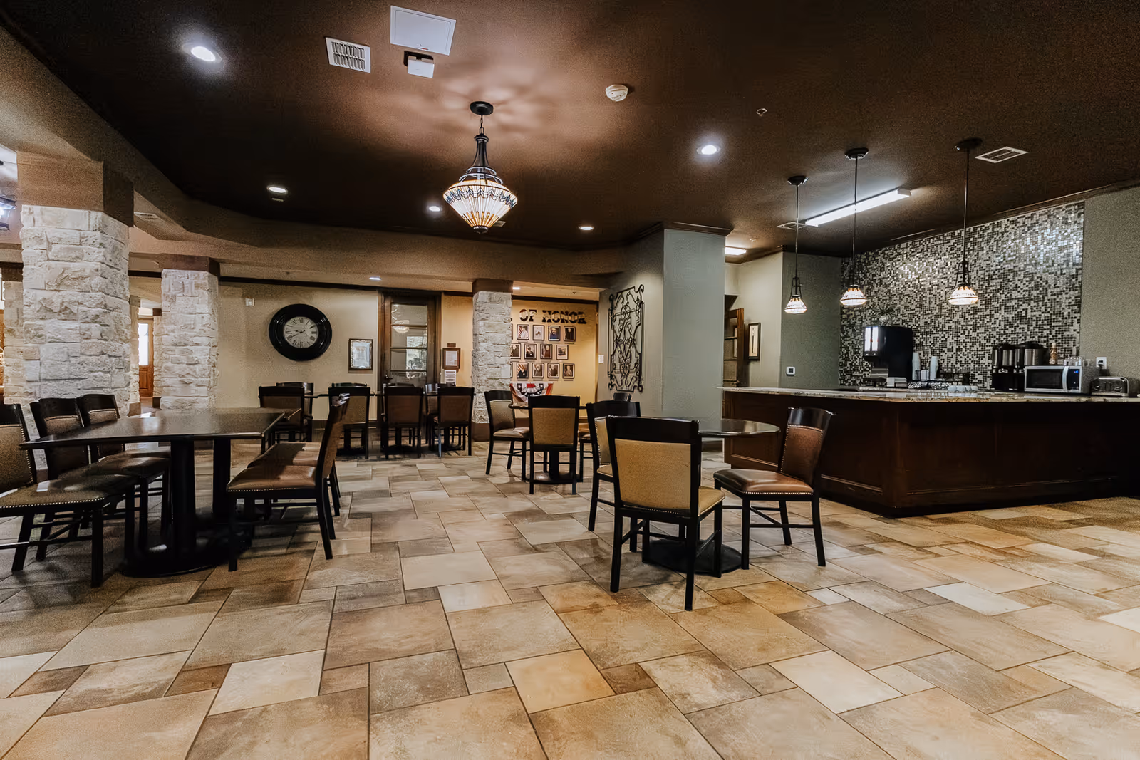 Spacious dining area with multiple tables and chairs, stone pillars, tiled floor, and a counter with pendant lights and kitchen appliances in the background.