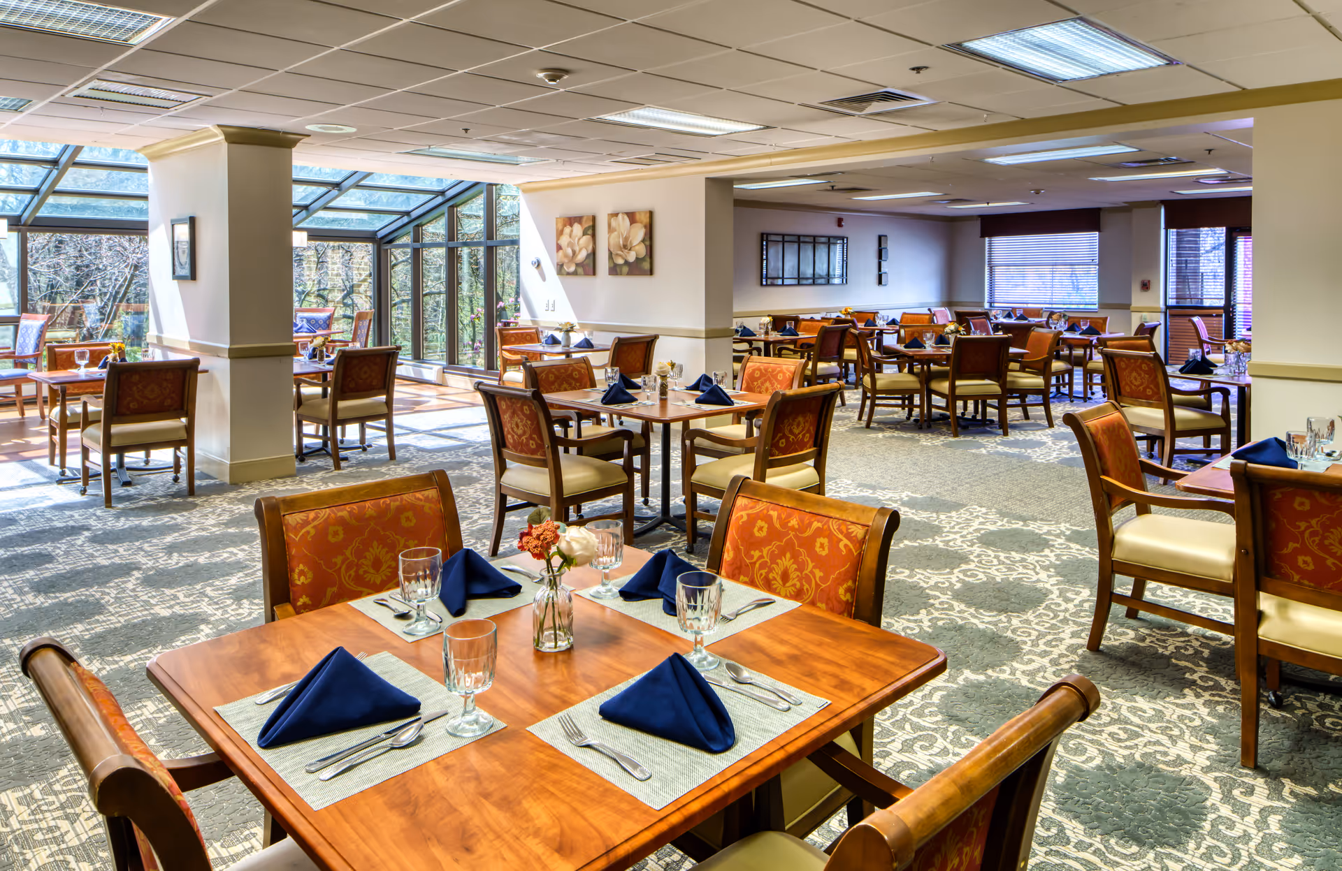 Sunlit dining room with multiple wooden tables set with blue napkins, glassware, and small floral centerpieces.