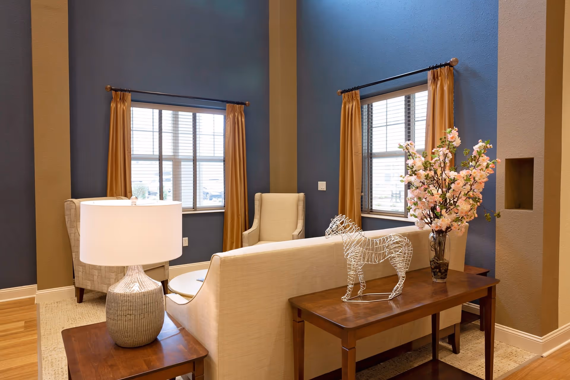 A cozy living room area with beige upholstered chairs and sofa, a wooden side table with a white lamp, and another wooden table with a decorative wire horse sculpture and a vase of pink flowers. The walls are painted blue with tan accents, and two windows with golden curtains let in natural light.