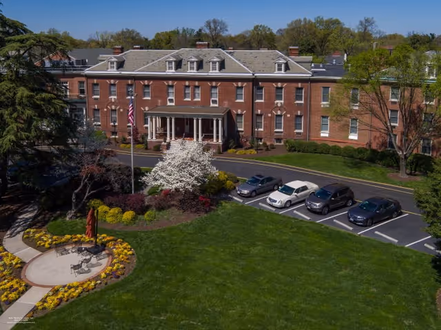 Front facade of a multi-story brick senior living building with a circular driveway, landscaped lawn, and parked cars.