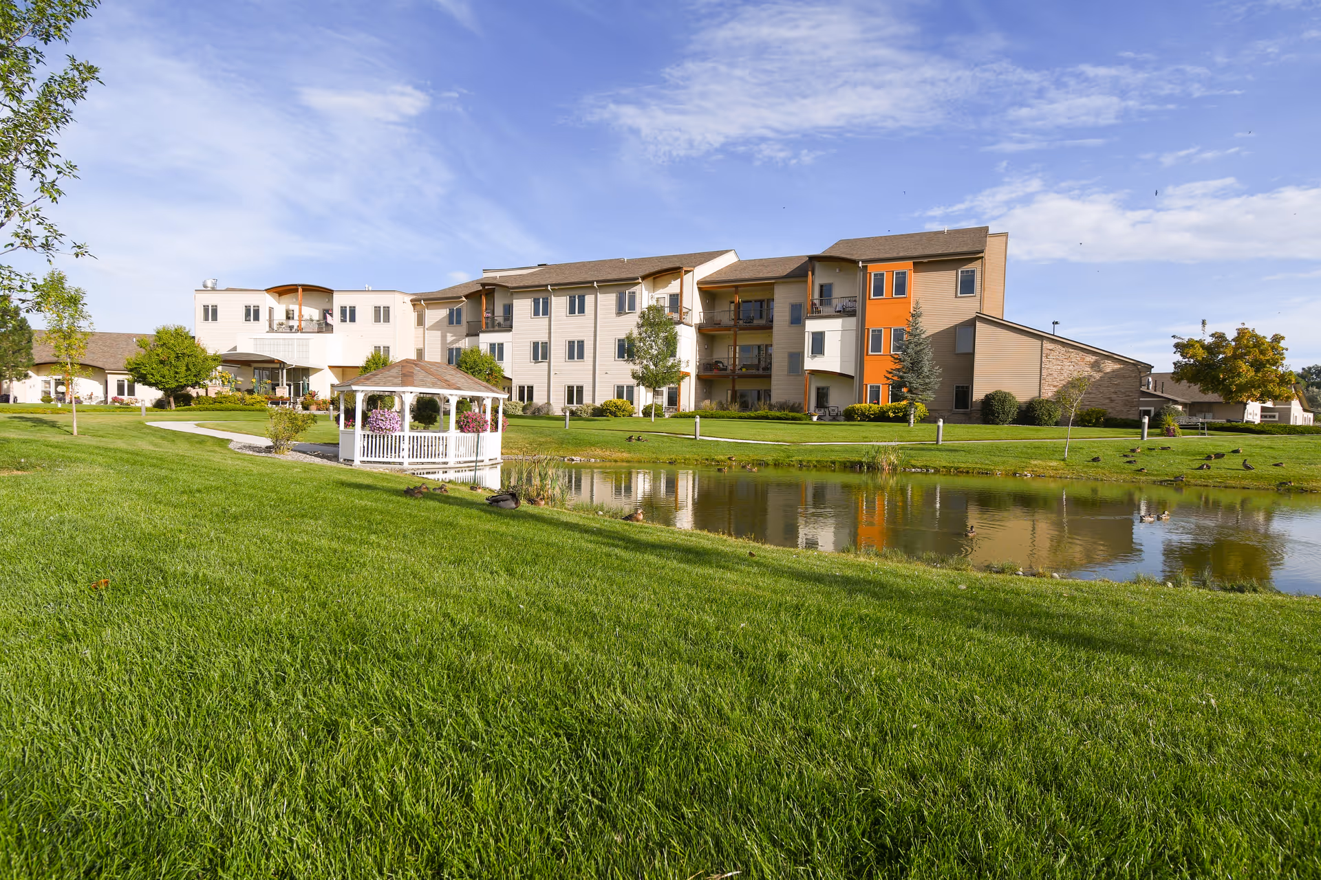 A senior living facility with a large green lawn, a small pond, and a white gazebo decorated with hanging flower baskets. The building is multi-story with balconies and a mix of beige and orange exterior walls under a partly cloudy blue sky.
