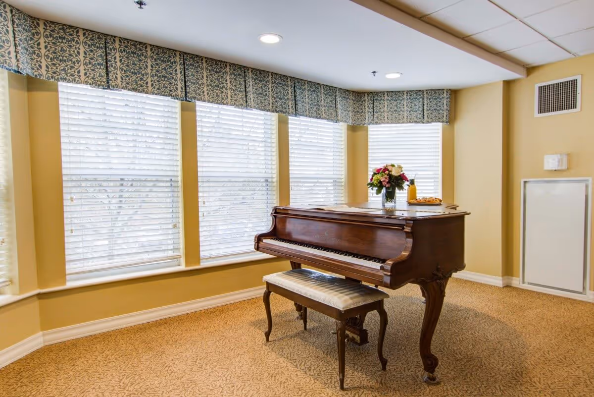 A wooden grand piano with a matching cushioned bench is placed in a carpeted room with beige walls. The piano has a vase of flowers and a plate with a bottle on top. Behind the piano, there are large windows with white blinds and patterned valances.