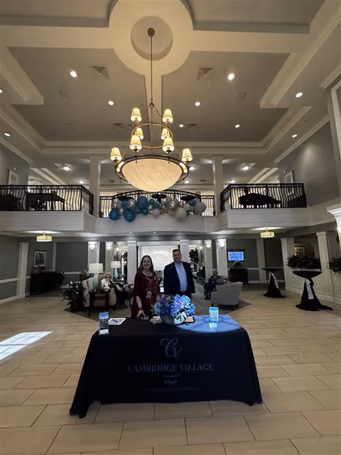 Spacious and elegant lobby area of Cambridge Village of Apex with high ceilings, a large chandelier, and a balcony decorated with blue and silver balloons. Two people stand behind a table covered with a black tablecloth displaying the Cambridge Village logo and name. Comfortable seating and plants are arranged around the lobby.