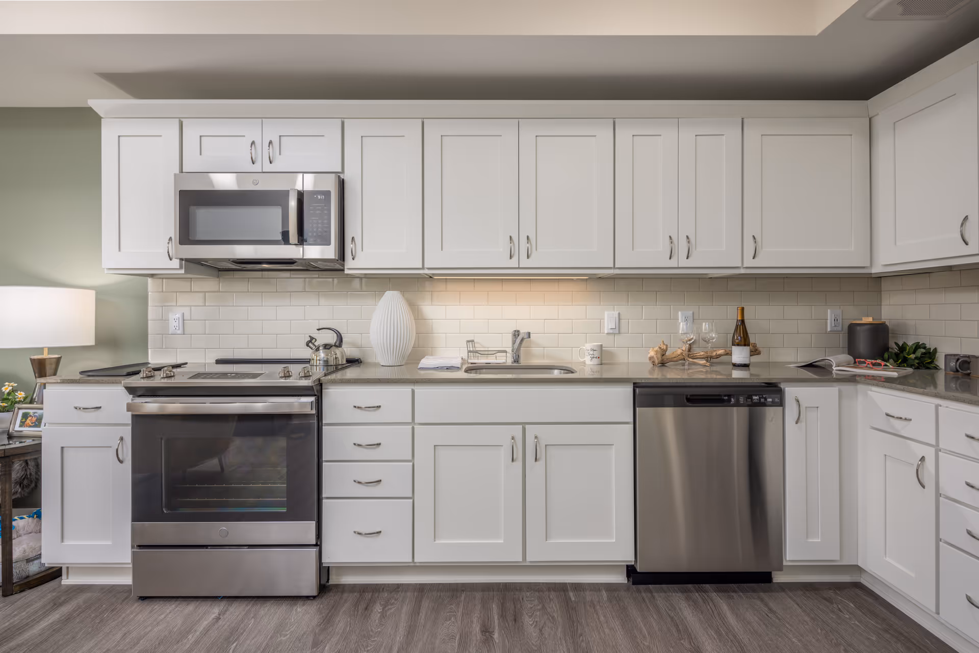 A modern kitchen with white cabinets and drawers, a stainless steel oven and microwave, a stainless steel dishwasher, a sink with a faucet, and a countertop decorated with a white vase, a tea kettle, a coffee mug, wine glasses, a bottle of wine, and some greenery. The backsplash is made of white subway tiles and the floor has a wood-like finish.