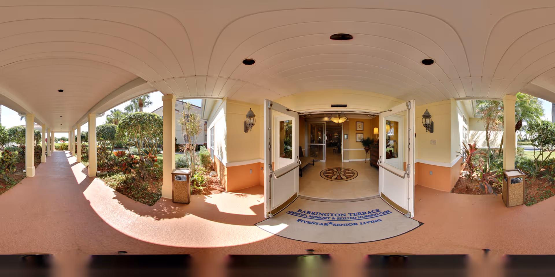 Covered entrance walkway leading to open double doors of Barrington Terrace senior living with a welcome mat and surrounding landscaping.