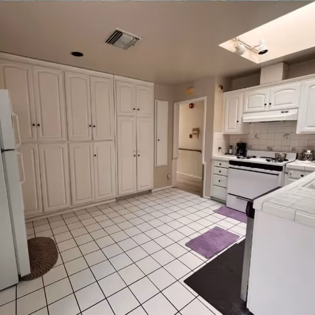 A clean kitchen with white tiled floor and white cabinetry. There is a white refrigerator on the left, a white stove with a range hood on the right, and multiple cabinets for storage. Two purple floor mats are placed near the stove and sink area. The kitchen has a skylight and a doorway leading to another room.