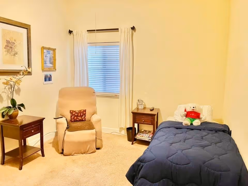 Small bedroom with a single bed topped by a blue comforter and teddy bear, a beige recliner, two wooden nightstands and a window with white curtains.