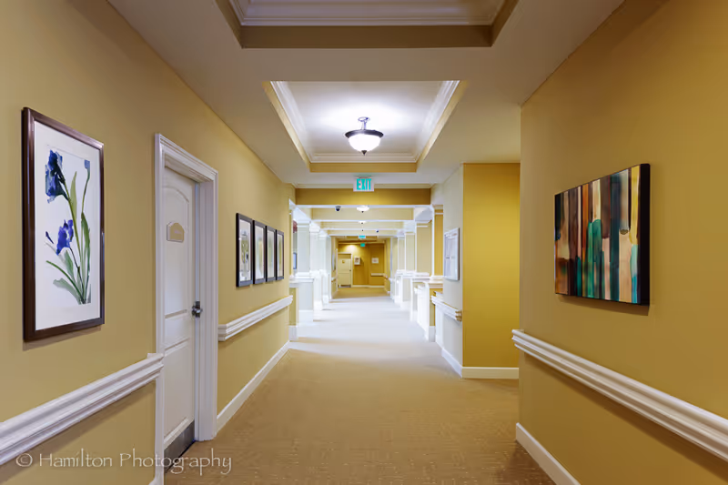 A well-lit hallway in a senior living facility with beige walls and carpeted floor. The hallway features white trim and molding, several framed artworks on the walls, and multiple doors along the corridor. Ceiling lights illuminate the space, and an exit sign is visible at the far end.