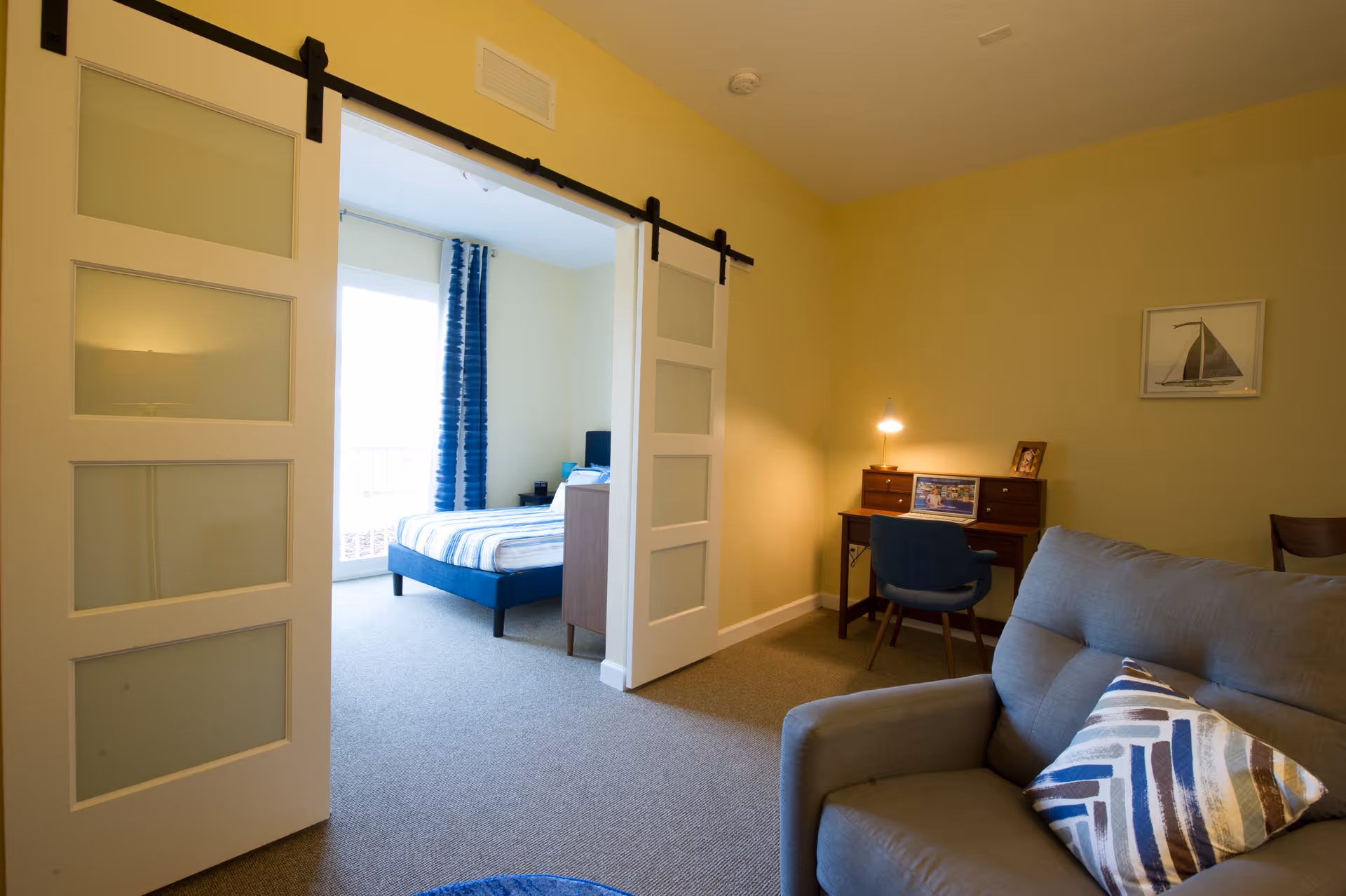 Interior view of a senior living facility room at Zon Beachside showing a living area with a gray armchair and a decorative pillow, a small wooden desk with a blue chair and a lit desk lamp, and a bedroom visible through sliding barn doors with a bed covered in blue and white striped bedding and blue curtains on the window.