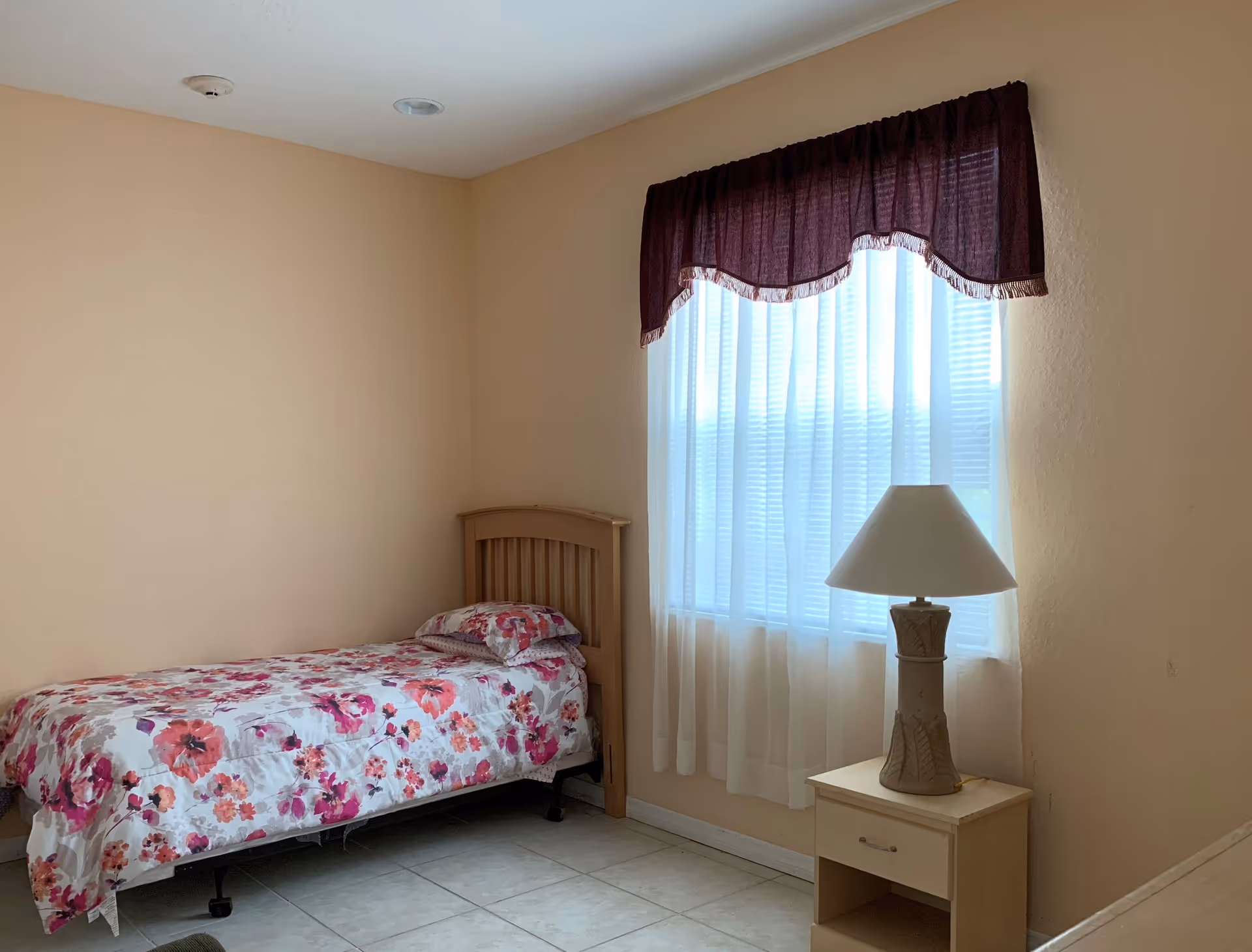 A small bedroom with single bed, floral bedding, nightstand, lamp, and window.