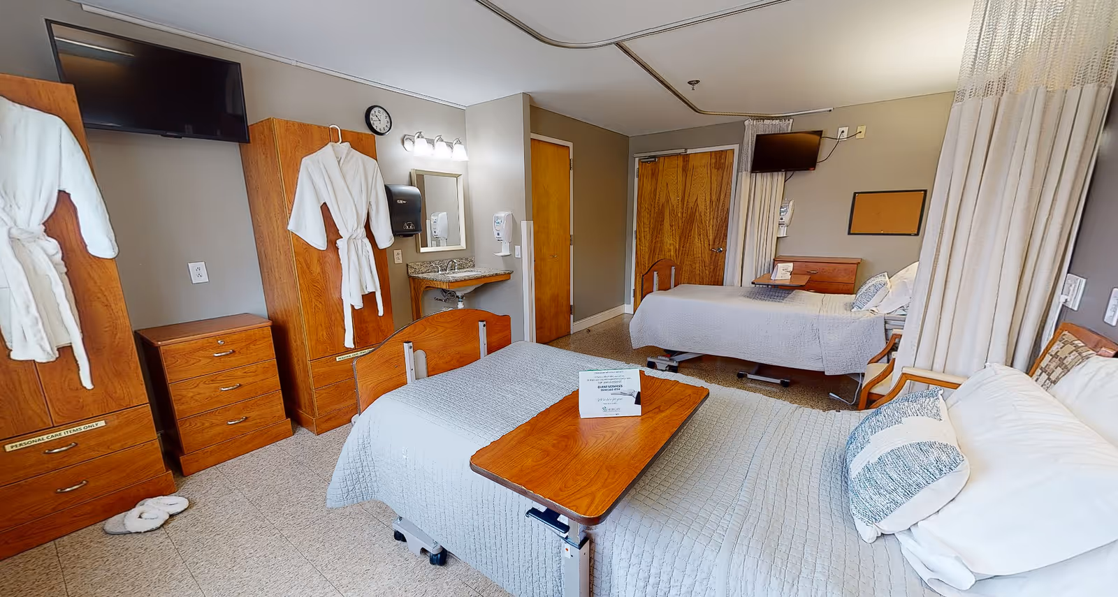 A shared patient room in a healthcare facility with two hospital beds, each with gray bedding and pillows. The room features wooden furniture including dressers and wardrobes, two wall-mounted flat-screen TVs, a small sink with a mirror and light above it, and two white robes hanging on the wardrobes. There is a privacy curtain partially drawn between the beds.