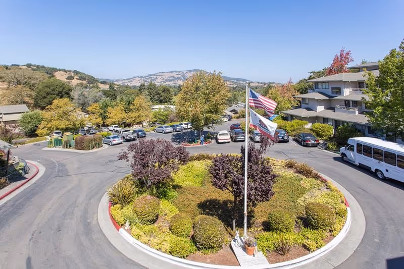 A circular driveway with a landscaped roundabout featuring various shrubs and small trees, three flagpoles with American and state flags, parked cars around the driveway, a white shuttle bus, and a multi-story building surrounded by trees and hills in the background under a clear sky.