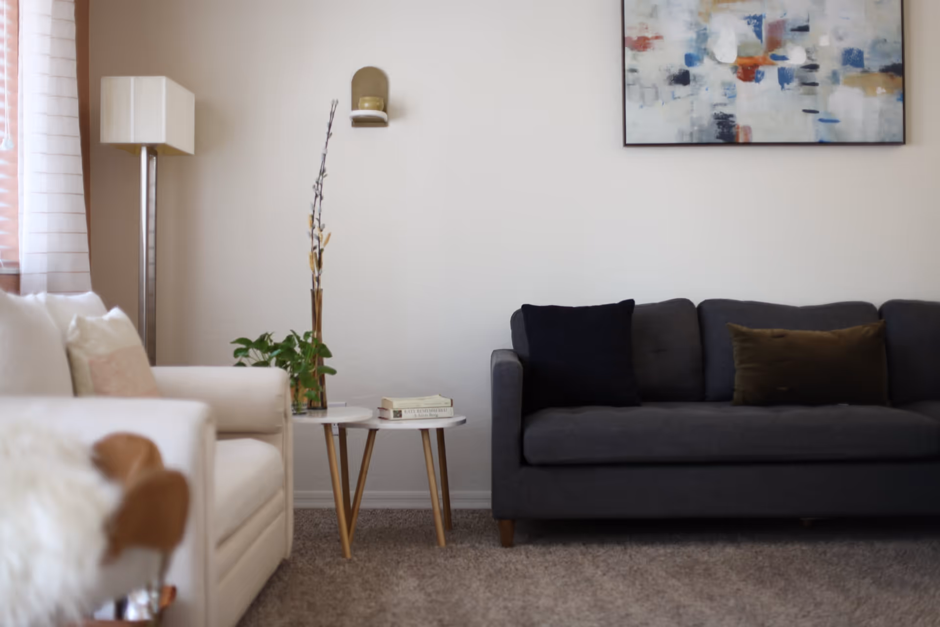 A cozy living room with a dark gray sofa adorned with two cushions, a white armchair with pillows, a small round side table holding books and a vase with dried branches, a floor lamp, and an abstract painting on the wall.