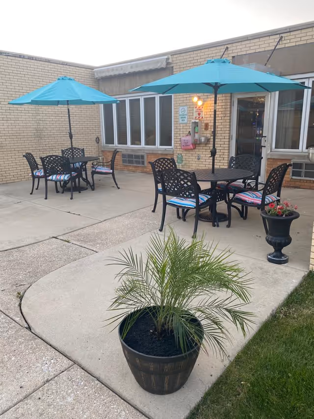 Outdoor patio area with two black metal tables, each surrounded by four black metal chairs with striped cushions. Each table has a large turquoise umbrella providing shade. The patio is adjacent to a brick building with several windows and a door. There are two potted plants, one with green foliage in the foreground and another with flowers near the building.