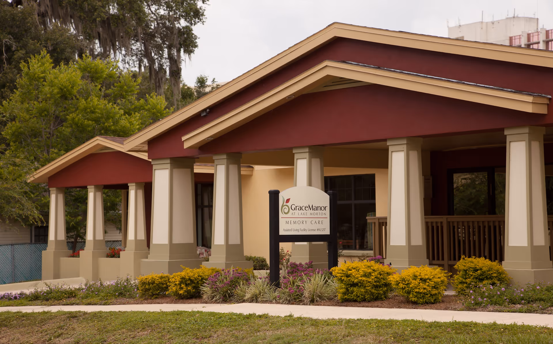 Exterior view of Grace Manor at Lake Morton, a memory care assisted living facility, showing a building with beige and maroon colors, columns supporting a covered porch, and landscaped bushes and flowers in front.