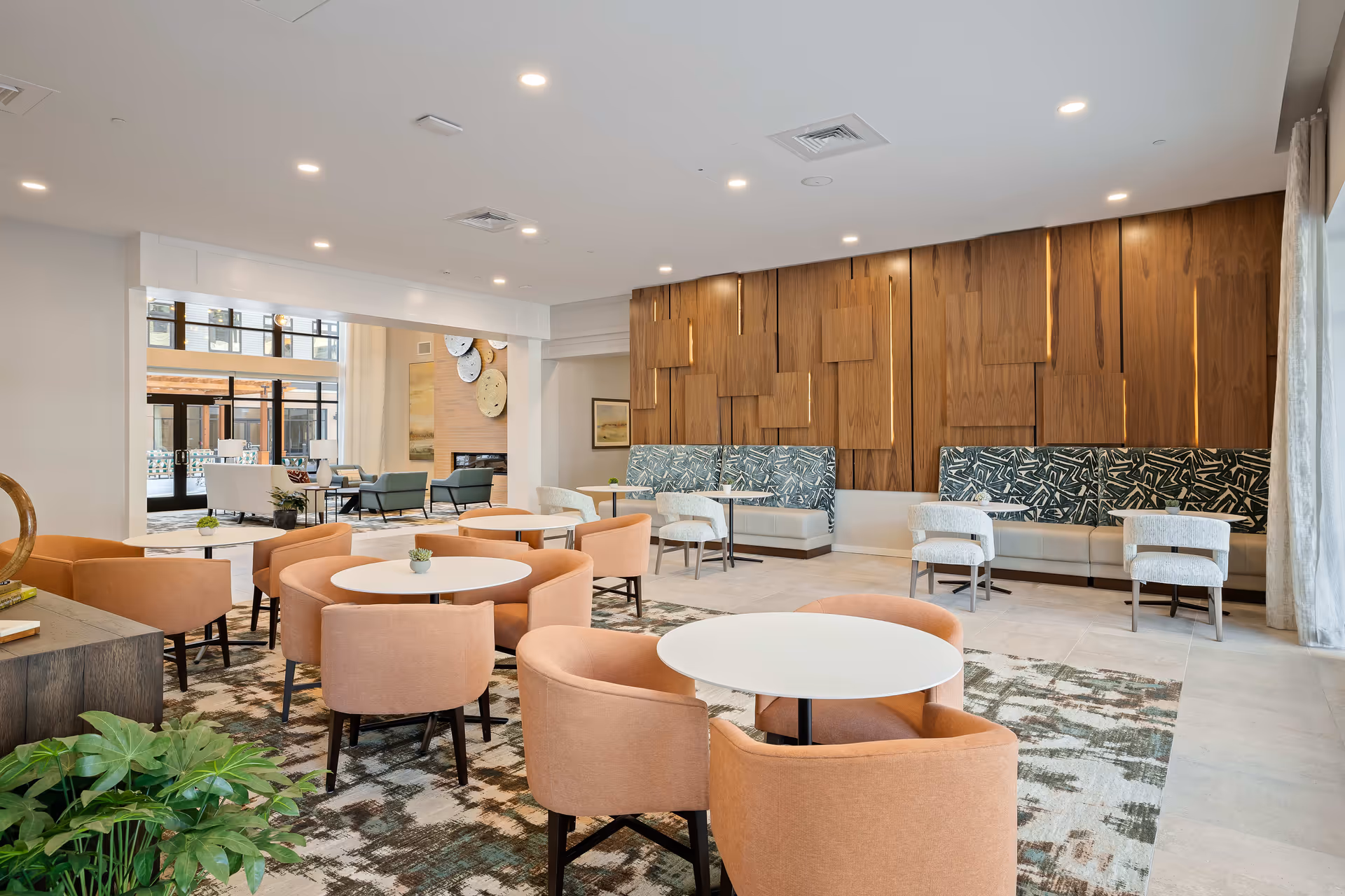 A modern and spacious common area with round white tables surrounded by peach-colored chairs. The back wall features decorative wooden panels with built-in lighting and patterned cushioned bench seating. The room is well-lit with recessed ceiling lights and has a large window with sheer curtains on the right side. In the background, there is another seating area with armchairs and a fireplace, visible through a large open doorway.