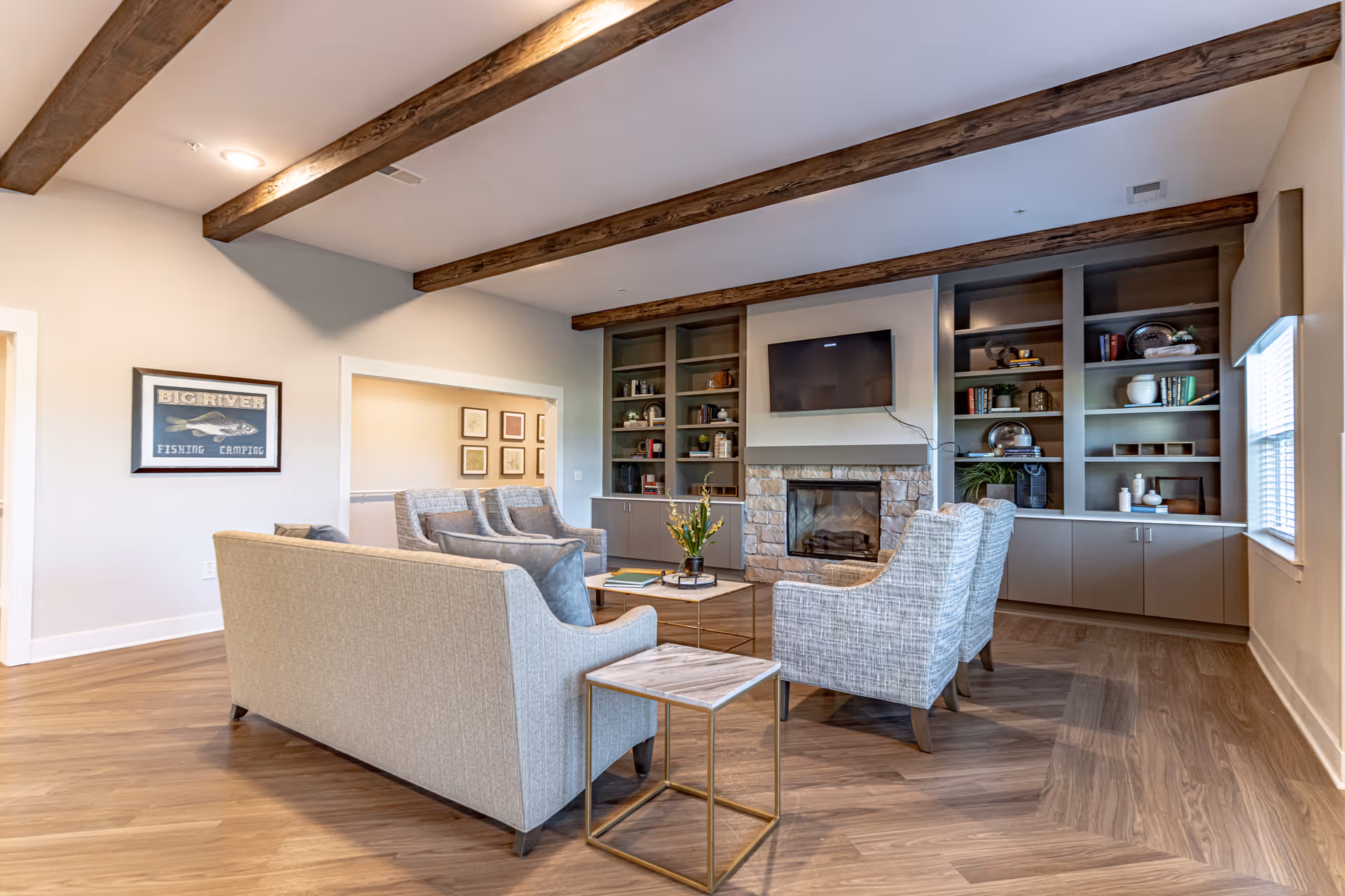 A cozy living room with wooden ceiling beams, a stone fireplace, a wall-mounted TV, built-in shelves filled with decorative items and books, and comfortable seating including a sofa and armchairs arranged around a coffee table.