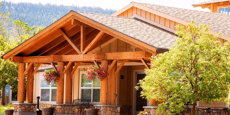 Exterior view of a senior living facility with a wooden porch structure supported by large timber beams. Hanging flower baskets decorate the porch area, and there is a small seating area with a bench. A tree with green leaves is visible on the right side, and mountains can be seen in the background under a clear sky.