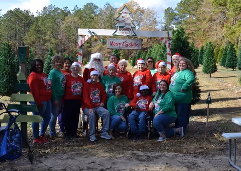 A group of people posing outdoors in a Christmas tree farm setting. They are wearing festive holiday sweaters and Santa hats, standing and sitting in front of a wooden arch decorated with candy cane stripes and a sign that reads 'Merry Christmas' and 'Fresh Cut Christmas Trees For Sale'. Trees and a wooded area are visible in the background.