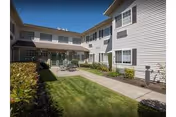 Outdoor courtyard with green lawn, paved walkway, and patio seating between two-story white-siding buildings under a blue sky.