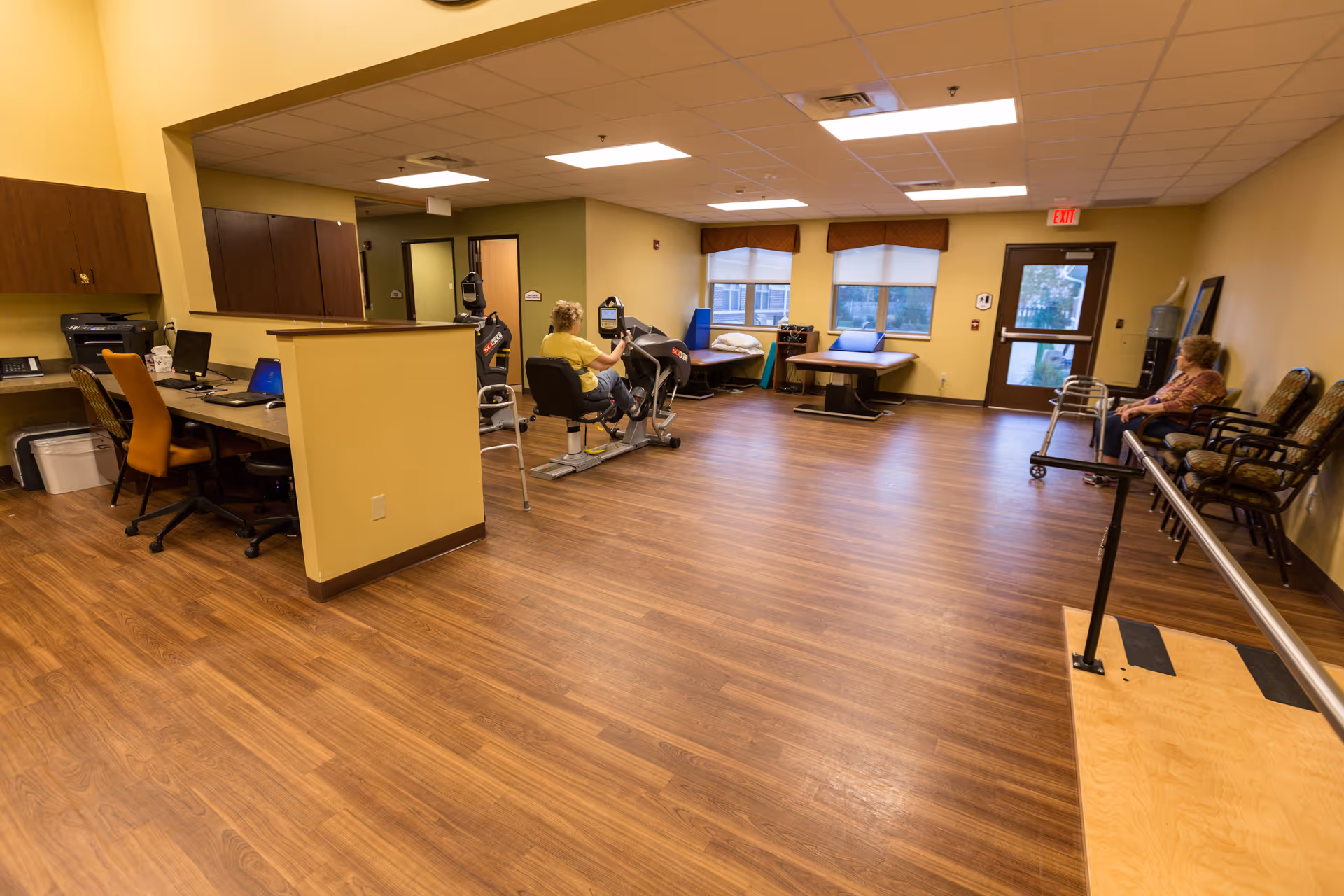A spacious room with wooden flooring in a senior living facility. The room contains exercise equipment with a person using a stationary bike, a therapy table near the windows, a desk area with computers and chairs, and a few chairs along the wall where another person is seated. The walls are painted yellow and there is a door leading outside.