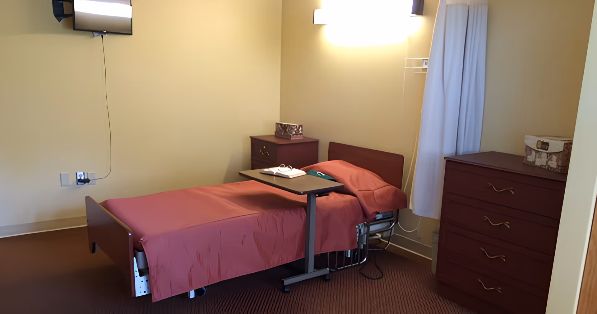 A small senior living bedroom with a single hospital-style bed covered in a red blanket, an overbed table, dresser, nightstand, wall-mounted TV, and a curtained window.