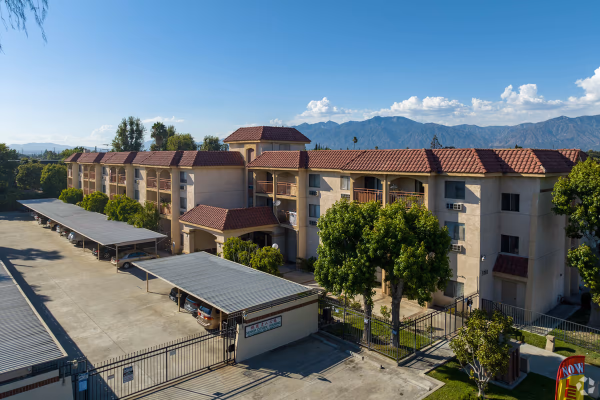 Three-story apartment building with red tile roofs, covered parking, a gated entrance, and mountains in the background.