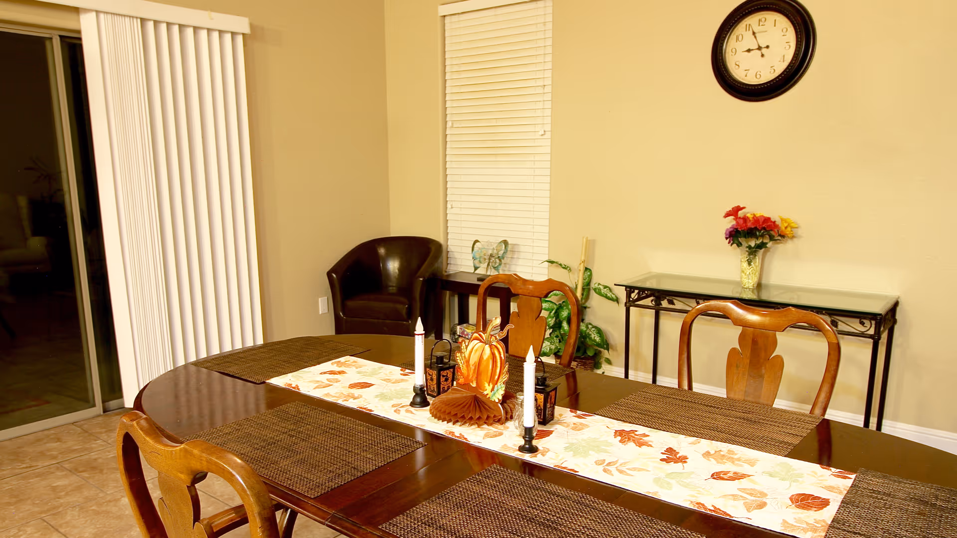 A dining room with a dark wooden table set with four brown woven placemats and a decorative autumn-themed table runner featuring candles and a pumpkin centerpiece. Behind the table are three wooden chairs, a small black console table with a vase of colorful flowers, a wall clock, a potted plant, and a brown leather armchair near a window with white blinds. Sliding glass doors with vertical blinds are visible on the left side.