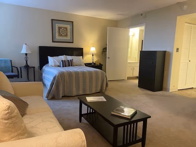 Spacious studio bedroom with a bed, sofa, coffee table, dresser, and an open doorway to a bathroom.