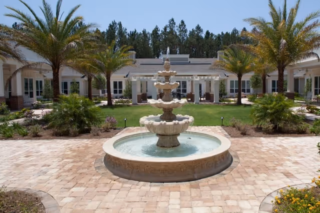 A courtyard with a multi-tiered stone fountain in the center, surrounded by palm trees, green grass, and a paved walkway. The courtyard is enclosed by a single-story building with large windows and a covered patio area. Tall pine trees are visible in the background under a clear blue sky.