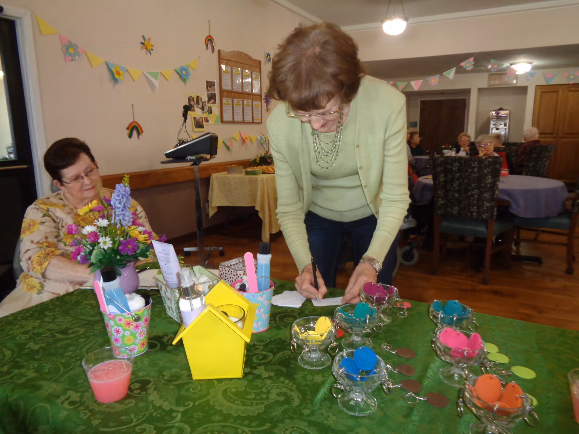 A group of elderly women in a decorated communal dining room, one woman leaning over a table covered with colorful craft supplies and decorations.